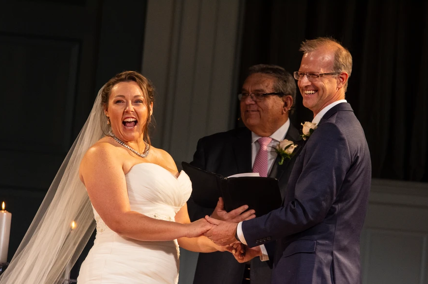 A bride is happily surprised as she holds hands with the groom who’s grinning with the officiant behind them.