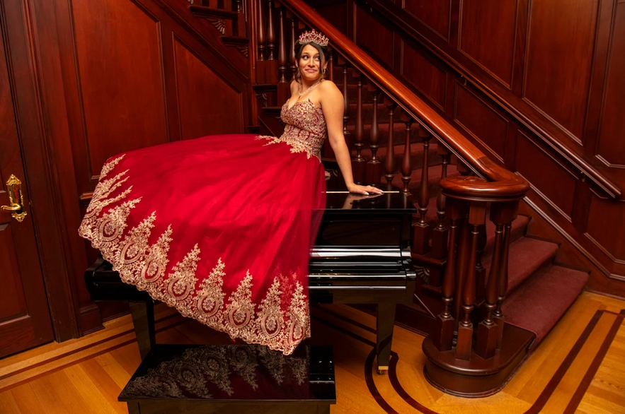 A young girl posing on top of a piano wearing a red gown for her Quinceañera party.